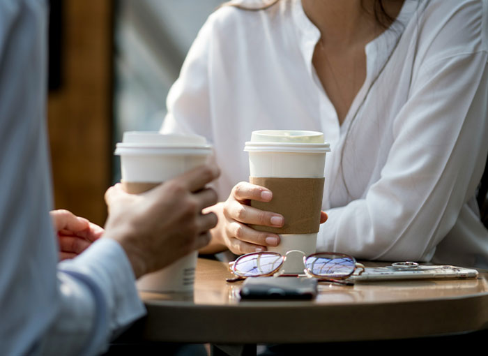 Two colleagues holding coffee cups at a table, one in a white shirt and another partially visible. Two colleagues holding coffee cups at a table, one in a white shirt and another partially visible.