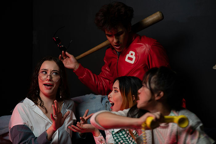 A woman with glasses animatedly talks among friends, while a man in a red jacket stands behind, creating a lively scene. A woman with glasses animatedly talks among friends, while a man in a red jacket stands behind, creating a lively scene.