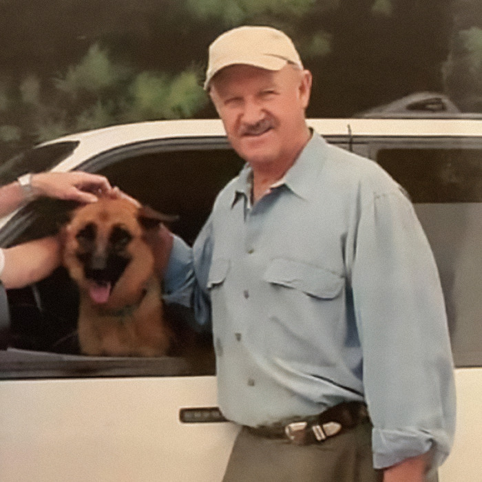 Gene Hackman in a blue shirt and cap stands by a car with a dog inside, smiling outdoors. Gene Hackman in a blue shirt and cap stands by a car with a dog inside, smiling outdoors.