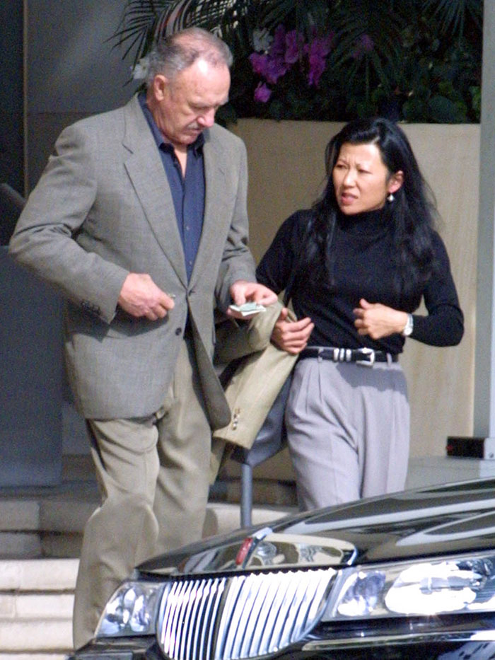 Elderly couple walking outdoors, man in suit, woman in black top, referencing "Last Photos of Gene Hackman and Betsy Arakawa. Elderly couple walking outdoors, man in suit, woman in black top, referencing "Last Photos of Gene Hackman and Betsy Arakawa.