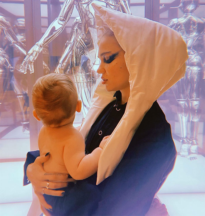 Grimes holding a baby, dressed in a unique outfit, against a futuristic background. Grimes holding a baby, dressed in a unique outfit, against a futuristic background.