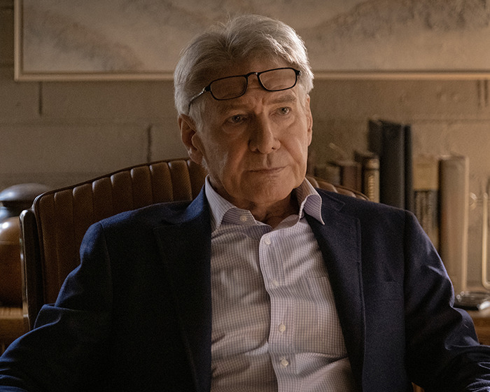 Harrison Ford seated with glasses atop head, wearing a suit and light shirt, thoughtful expression against bookshelf backdrop in a scene from "Shrinking. Harrison Ford seated with glasses atop head, wearing a suit and light shirt, thoughtful expression against bookshelf backdrop in a scene from "Shrinking.