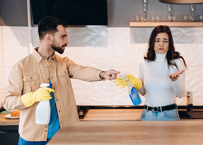 Husband and wife cleaning the kitchen, wearing gloves, engaged in a discussion about chores. Husband and wife cleaning the kitchen, wearing gloves, engaged in a discussion about chores.