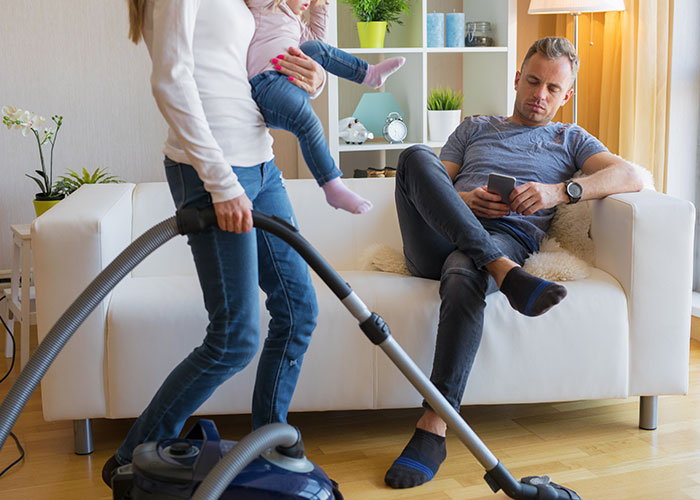 Husband on couch using phone while wife vacuums holding child, highlighting "unhelpful" dynamic. Husband on couch using phone while wife vacuums holding child, highlighting "unhelpful" dynamic.