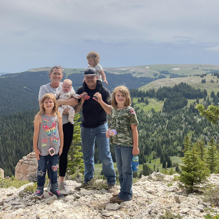 Family poses outdoors on a mountain with trees in the background; father stands with four daughters and mother. Family poses outdoors on a mountain with trees in the background; father stands with four daughters and mother.