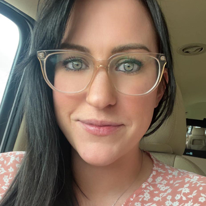 A woman with dark hair and glasses sits in a car, wearing a pink floral top. A woman with dark hair and glasses sits in a car, wearing a pink floral top.