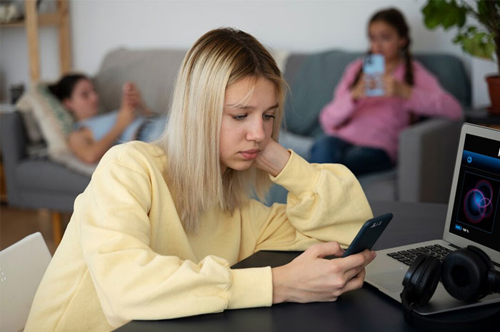 Teen girl looking at phone, appearing frustrated, while sitting near a laptop, wearing a yellow sweater in a living room. Teen girl looking at phone, appearing frustrated, while sitting near a laptop, wearing a yellow sweater in a living room.