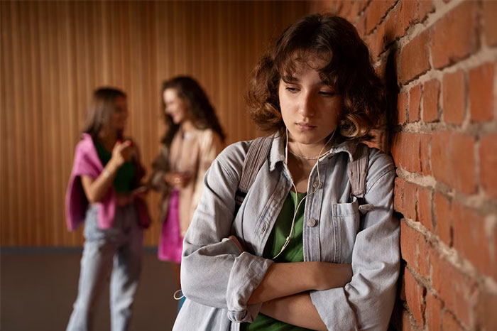 Teen girl upset by brick wall, classmates in background, dealing with name issues. Teen girl upset by brick wall, classmates in background, dealing with name issues.