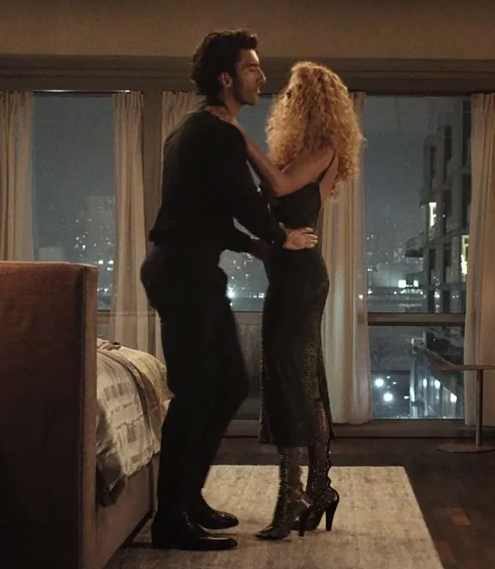 A couple in a dimly lit room, standing close, with city lights in the background, discussing "fat-shaming" accusations. A couple in a dimly lit room, standing close, with city lights in the background, discussing "fat-shaming" accusations.