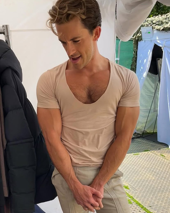 Jonathan Bailey showing toned muscles in a fitted beige shirt, standing outside with a jacket hung on a rack. Jonathan Bailey showing toned muscles in a fitted beige shirt, standing outside with a jacket hung on a rack.