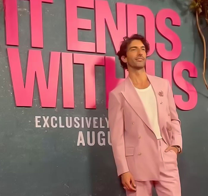 Man in a pink suit poses at "It Ends With Us" event, against a promotional background. Man in a pink suit poses at "It Ends With Us" event, against a promotional background.
