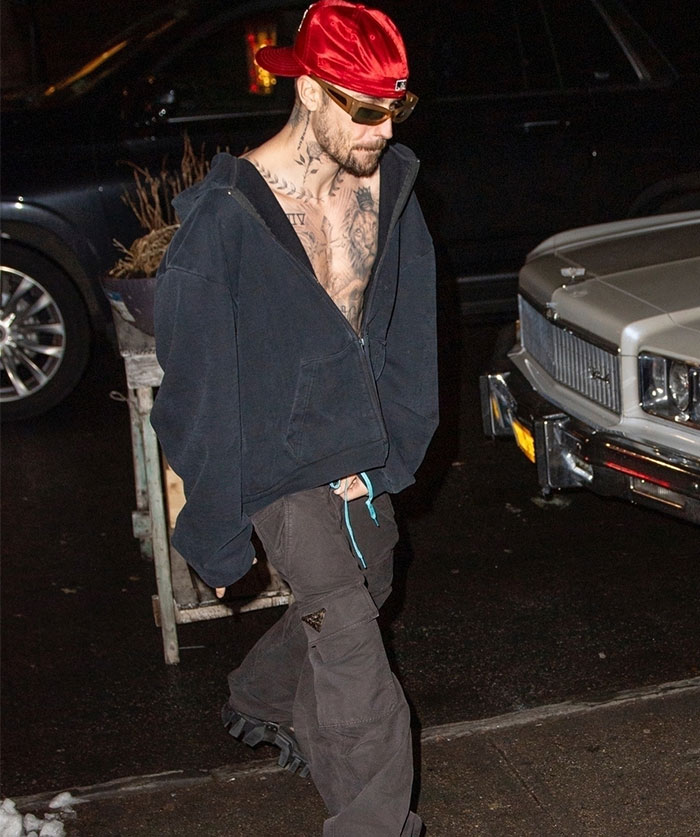Man in urban attire with tattoos walks by a car at night, wearing a red cap and sunglasses, capturing a casual moment. Man in urban attire with tattoos walks by a car at night, wearing a red cap and sunglasses, capturing a casual moment.