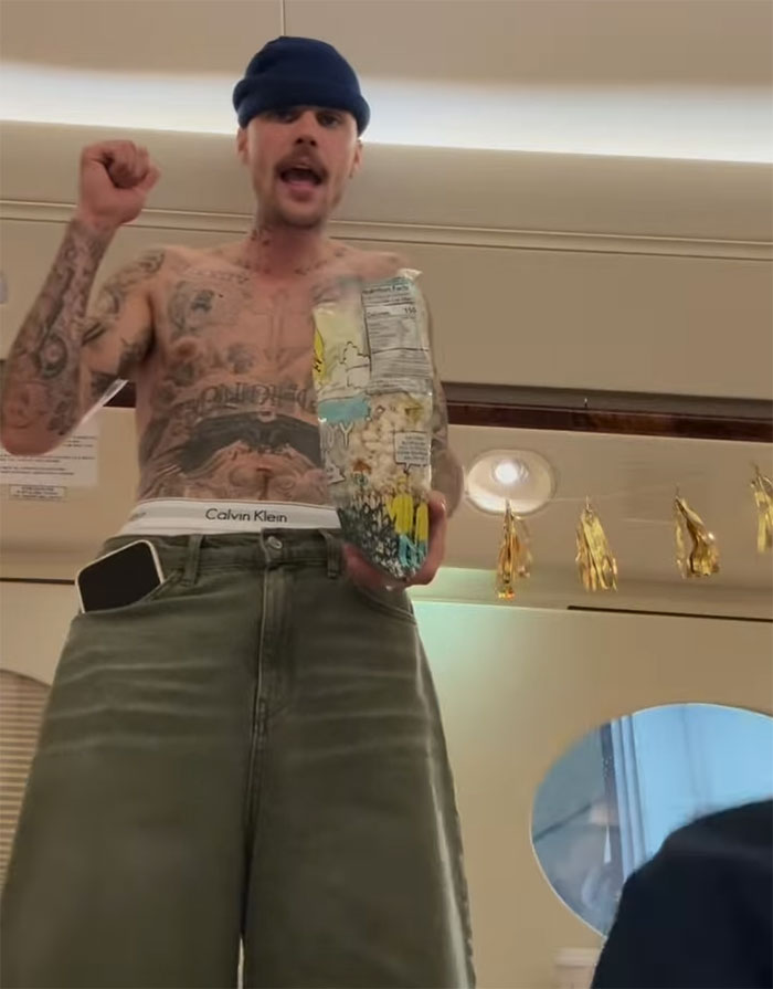 Shirtless man with tattoos holding a bag of snacks indoors, wearing a blue beanie and jeans, with golden decorations overhead. Shirtless man with tattoos holding a bag of snacks indoors, wearing a blue beanie and jeans, with golden decorations overhead.