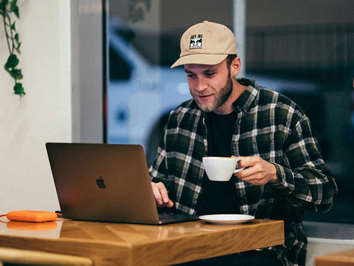 Man in a plaid shirt and hat, seated with a laptop and coffee, symbolizing taking time for himself. Man in a plaid shirt and hat, seated with a laptop and coffee, symbolizing taking time for himself.