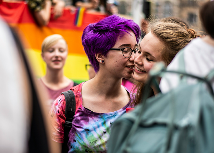 Two people embrace at a pride parade, celebrating LGBTQ+ identity with a rainbow flag in the background. Two people embrace at a pride parade, celebrating LGBTQ+ identity with a rainbow flag in the background.