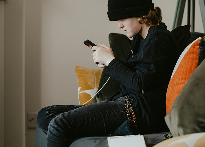 Teen in dark clothing sitting on a couch, focused on a smartphone, with pillows in the background. Teen in dark clothing sitting on a couch, focused on a smartphone, with pillows in the background.