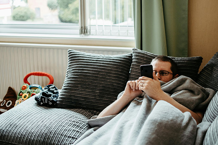 Man neglecting chores, lying on couch engrossed in phone, surrounded by unkempt living room with toys and pillows. Man neglecting chores, lying on couch engrossed in phone, surrounded by unkempt living room with toys and pillows.