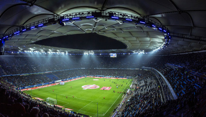 Wide-angle view of a brightly lit football stadium filled with spectators during an evening match. Wide-angle view of a brightly lit football stadium filled with spectators during an evening match.
