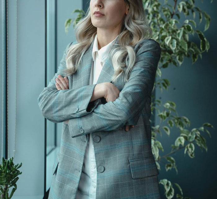 Woman in a plaid blazer stands with arms crossed, looking out of a window, contemplating something private on her laptop. Woman in a plaid blazer stands with arms crossed, looking out of a window, contemplating something private on her laptop.