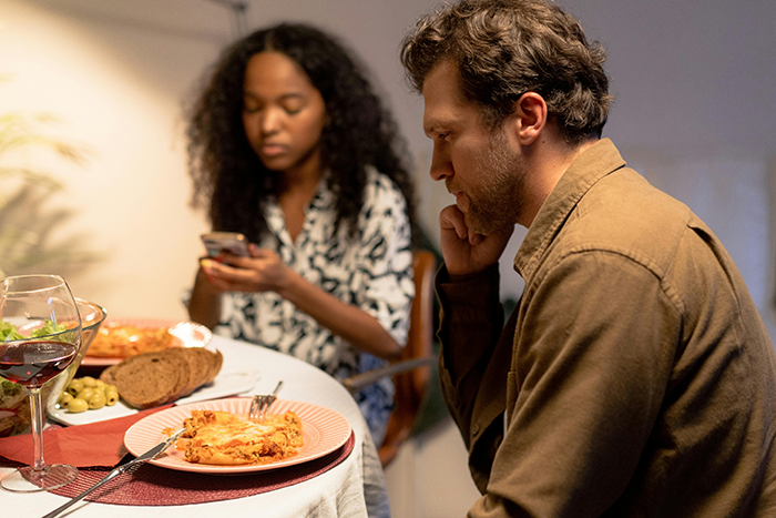A couple at dinner, the man looking at a lasagna plate, the woman checking her phone; lasagna and hot sauce theme. A couple at dinner, the man looking at a lasagna plate, the woman checking her phone; lasagna and hot sauce theme.