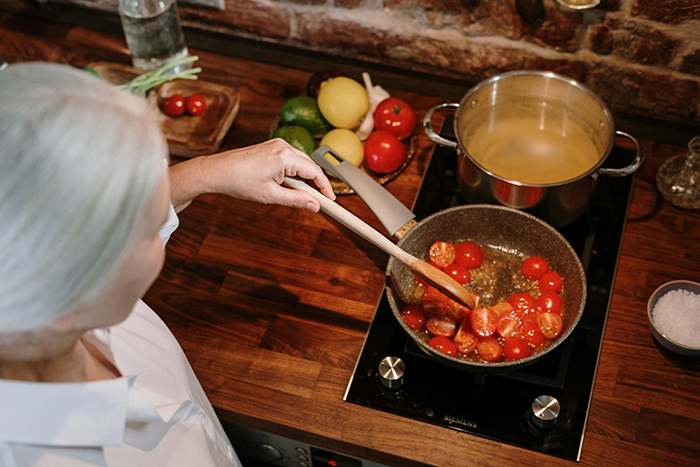 Older woman cooking lasagna ingredients on a stove, featuring cherry tomatoes and a large pot, with vegetables nearby. Older woman cooking lasagna ingredients on a stove, featuring cherry tomatoes and a large pot, with vegetables nearby.