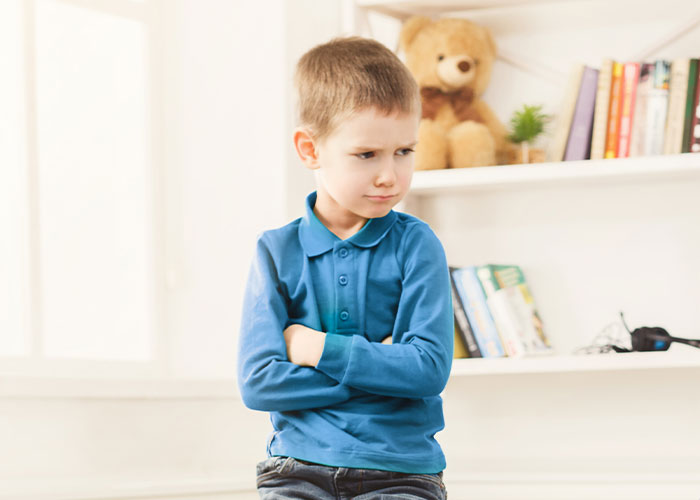Young boy in a blue shirt crossing arms, standing in a cozy room with a teddy bear and books on a shelf. Young boy in a blue shirt crossing arms, standing in a cozy room with a teddy bear and books on a shelf.