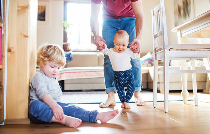 Parent helping baby walk while toddler sits nearby, showcasing sibling dynamics and parenting challenges. Parent helping baby walk while toddler sits nearby, showcasing sibling dynamics and parenting challenges.