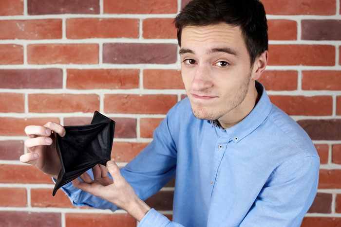 Man in a blue shirt showing an empty wallet, standing against a brick wall, symbolizing neighborhood loans struggle. Man in a blue shirt showing an empty wallet, standing against a brick wall, symbolizing neighborhood loans struggle.