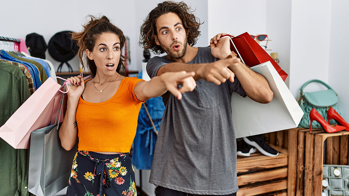 Two shoppers holding multiple thrift store finds while excitedly pointing at items in a shop filled with clothes and accessories