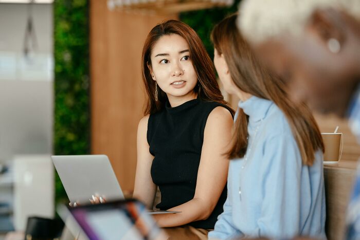 Two colleagues discussing work-struggles at a desk, one holding a laptop.