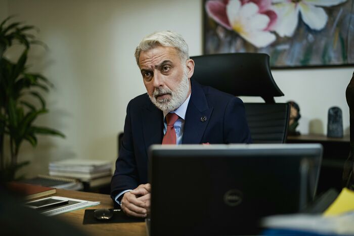 Man in an office, appearing thoughtful, seated at a desk, possibly facing work-struggles with a laptop and folders around.