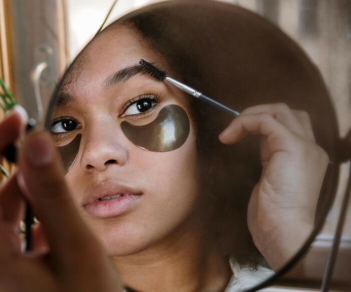 Person applying eye patches, reflecting on recent fashion trends people hate, using eyebrow brush.