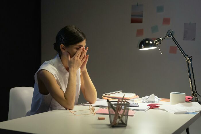 Person overwhelmed by work-struggles at a dimly lit desk, head in hands, surrounded by papers and a lamp.