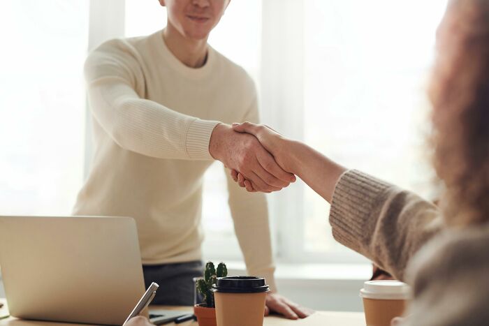Two people shaking hands in an office setting with coffee cups and a laptop nearby, illustrating workplace interaction.