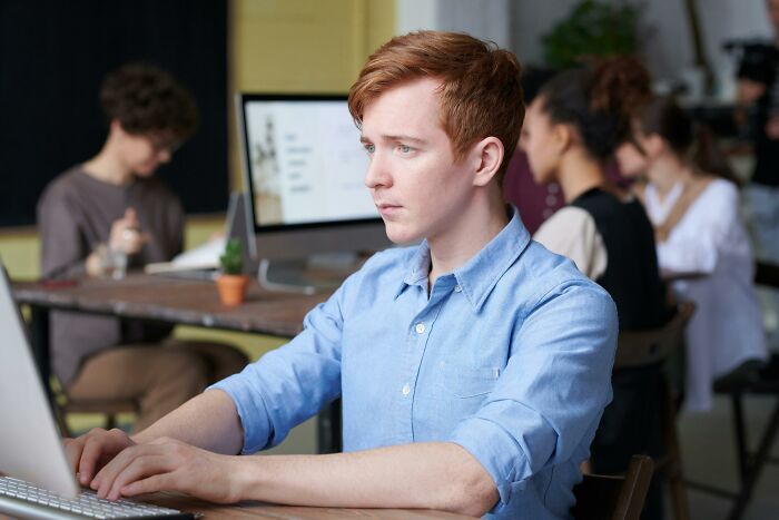 Person in a blue shirt facing a laptop, surrounded by colleagues at desks, highlighting work-struggles in a modern office.