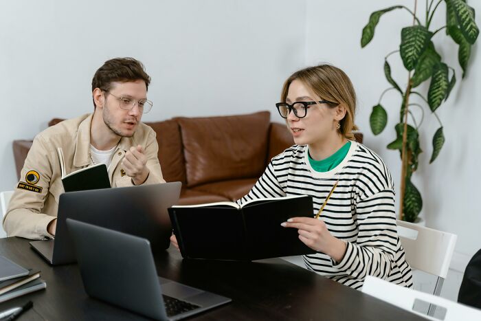 Two colleagues discussing work-struggles over a table with laptops and notebooks.