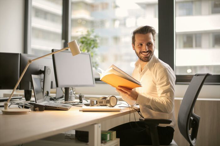 Person smiling at a desk, holding a book, with computers in an office setting, highlighting people lacking work skills.