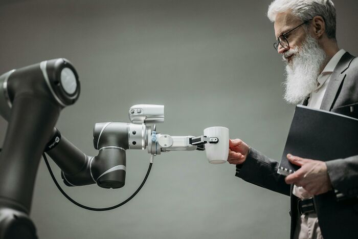 A robot arm hands a coffee mug to a man in a suit, illustrating jokes about office technology.