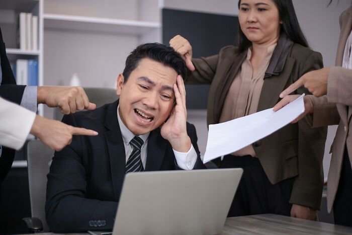 Stressed man at work surrounded by colleagues pointing fingers, highlighting lack of workplace skills.