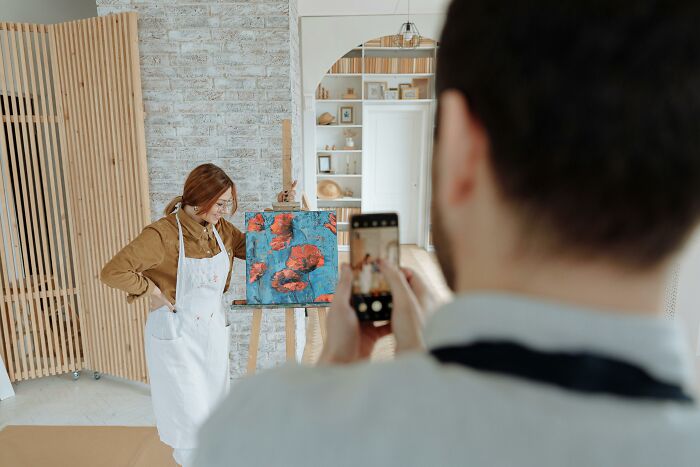 Woman in apron posing with painting while being photographed, highlighting social rules people ignore in personal space.