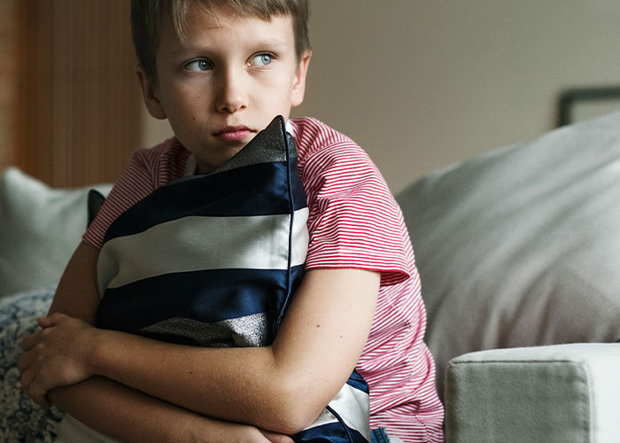 Young boy in a red striped shirt sitting on a couch, hugging a pillow and looking sad. Young boy in a red striped shirt sitting on a couch, hugging a pillow and looking sad.
