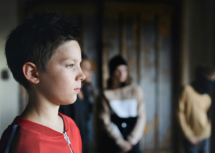 Young boy looking serious, wearing a red shirt, with blurred stepsiblings in the background during a family gathering. Young boy looking serious, wearing a red shirt, with blurred stepsiblings in the background during a family gathering.