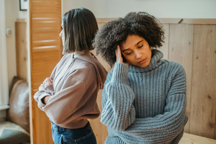 Two women standing back-to-back in a room, one looking frustrated; concept of freedom and family expectations. Two women standing back-to-back in a room, one looking frustrated; concept of freedom and family expectations.