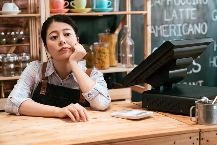 Restaurant worker covering shift, looking bored, sitting by a cash register, with coffee shop decor in the background. Restaurant worker covering shift, looking bored, sitting by a cash register, with coffee shop decor in the background.