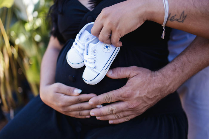 Pregnant woman holding baby shoes with returning husband’s hands on her belly. Pregnant woman holding baby shoes with returning husband’s hands on her belly.