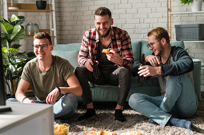 Three roommates laughing, sitting on the floor with snacks and drinks, enjoying a casual gathering. Three roommates laughing, sitting on the floor with snacks and drinks, enjoying a casual gathering.