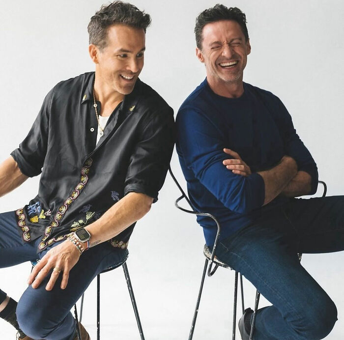 Two men laughing together while seated, wearing casual shirts and jeans, against a plain background. Two men laughing together while seated, wearing casual shirts and jeans, against a plain background.