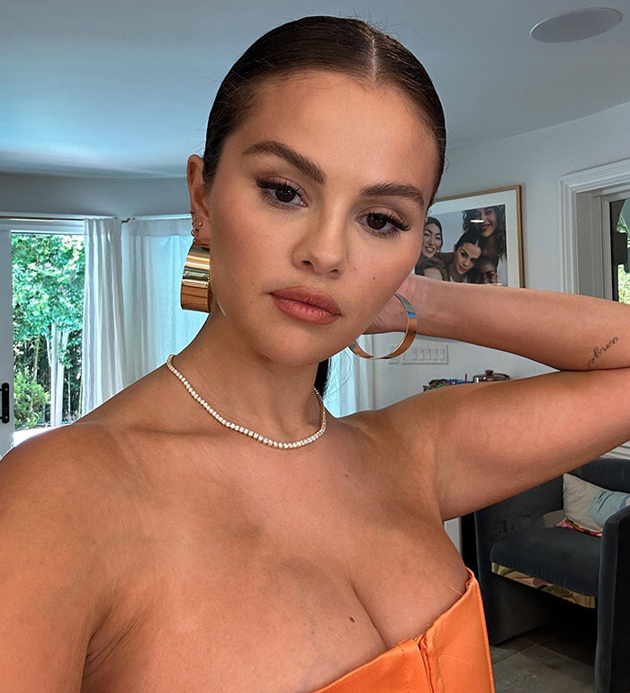 Selena Gomez showcasing weight loss, wearing a strapless orange dress and gold hoop earrings in a living room setting. Selena Gomez showcasing weight loss, wearing a strapless orange dress and gold hoop earrings in a living room setting.
