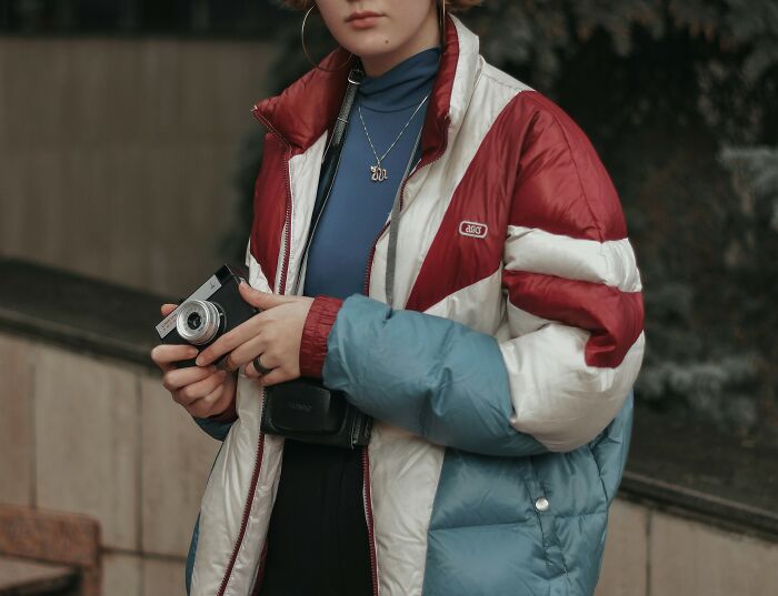 Person in a red and blue puffer jacket holding a vintage camera, showcasing recent fashion trends people hate.