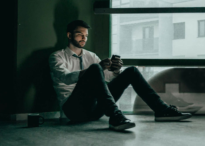 Man in a white shirt, sitting on the floor, looking at his phone with a serious expression. Man in a white shirt, sitting on the floor, looking at his phone with a serious expression.